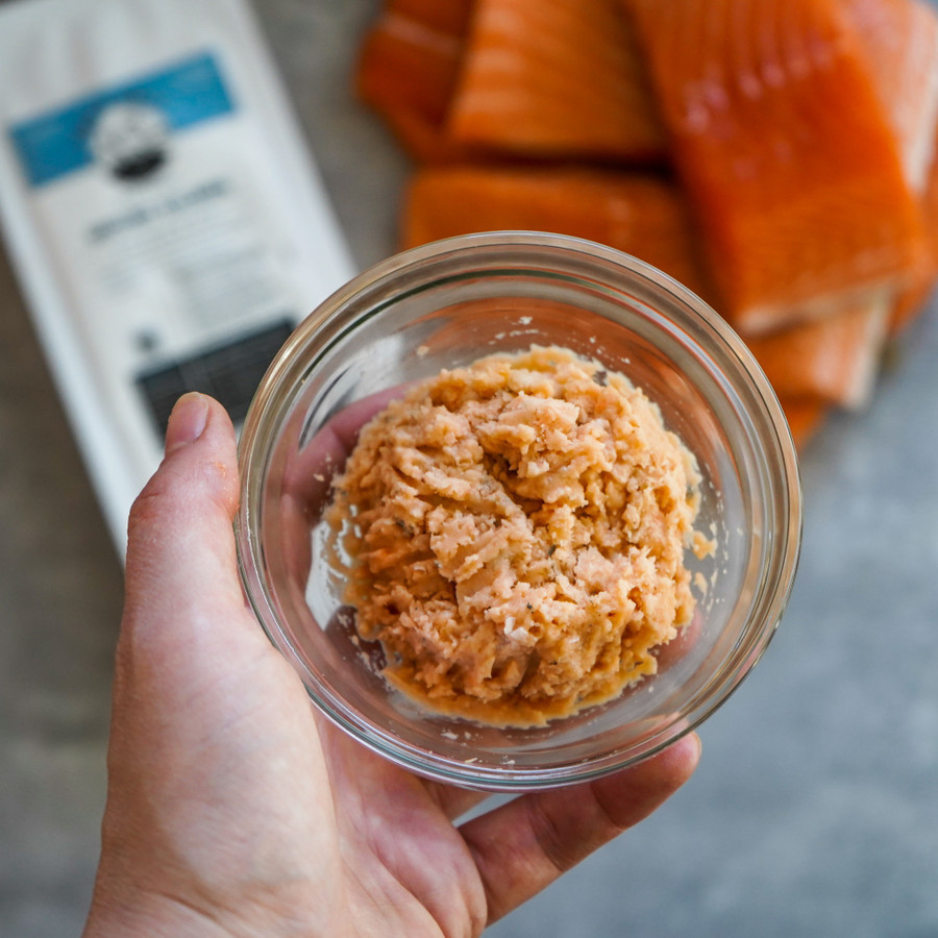 A hand holding a small glass bowl filled with ground salmon pet food, with salmon fillets and the product package blurred in the background on a kitchen counter.
