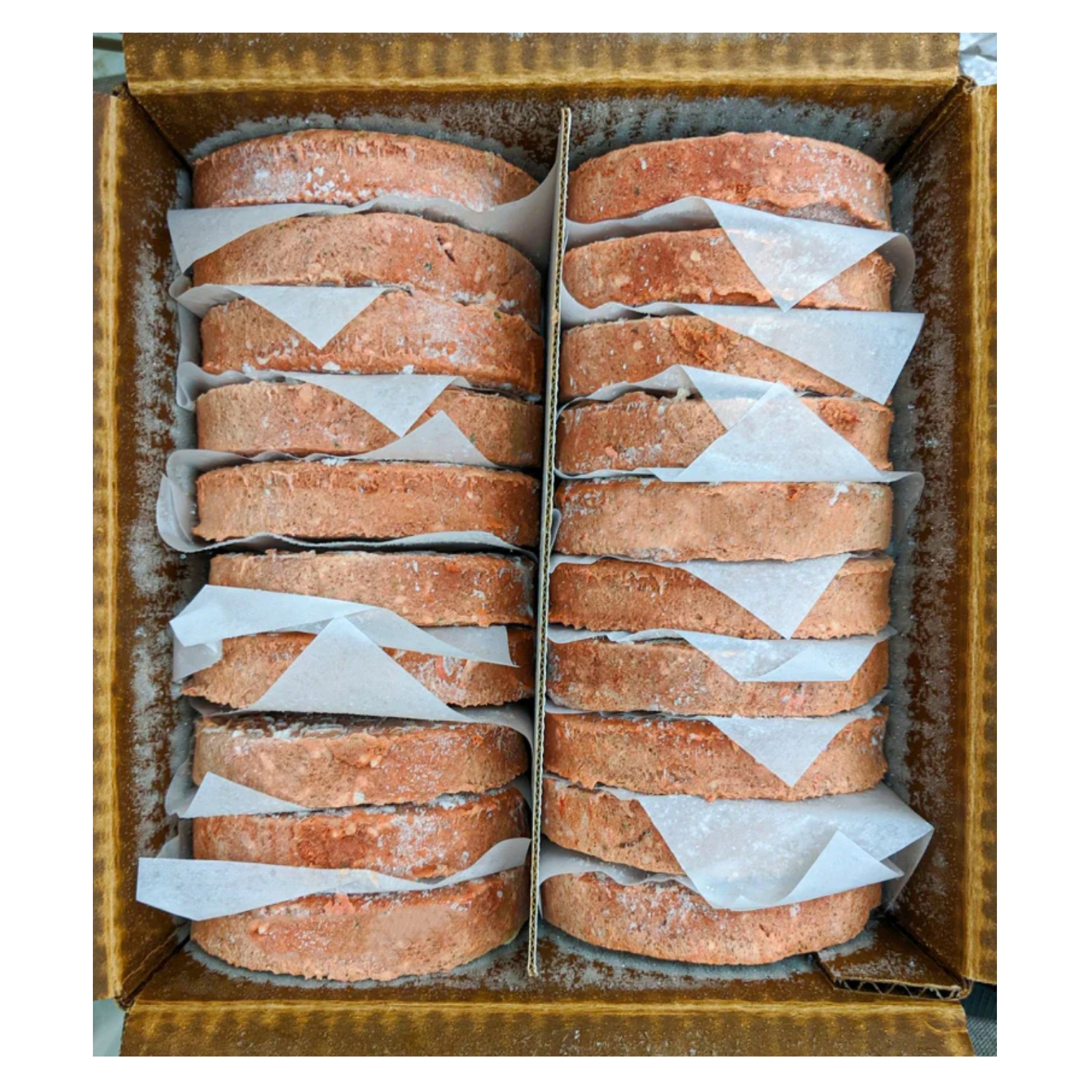 Top-down view of a cardboard box containing multiple frozen raw dog food patties stacked in rows and separated by sheets of white parchment paper. The patties are round, pinkish-brown in color, and lightly frosted.