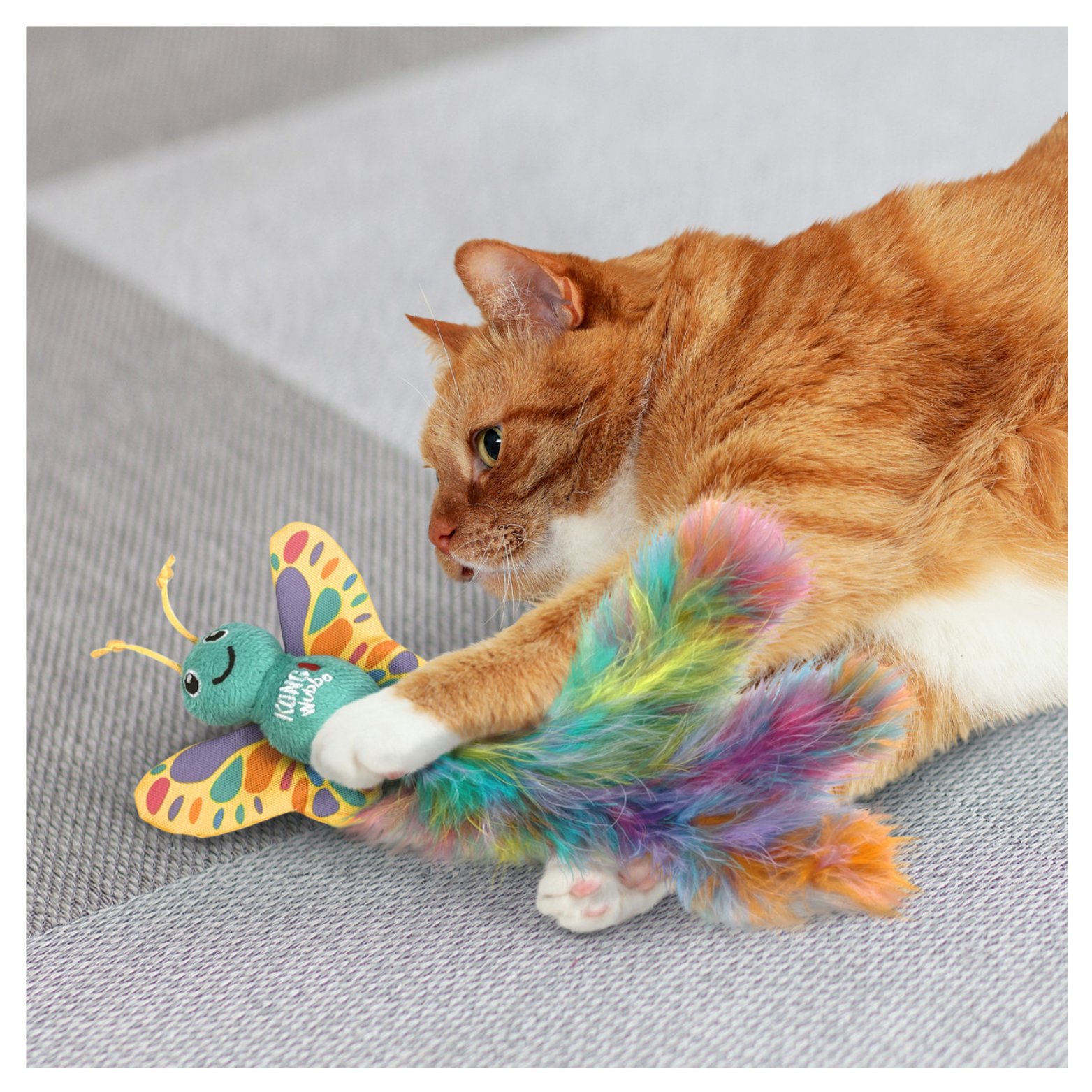 Orange tabby cat lying on a textured floor while holding and kicking the KONG Wubba Butterfly catnip toy, gripping the butterfly body and feather tail with its paws.