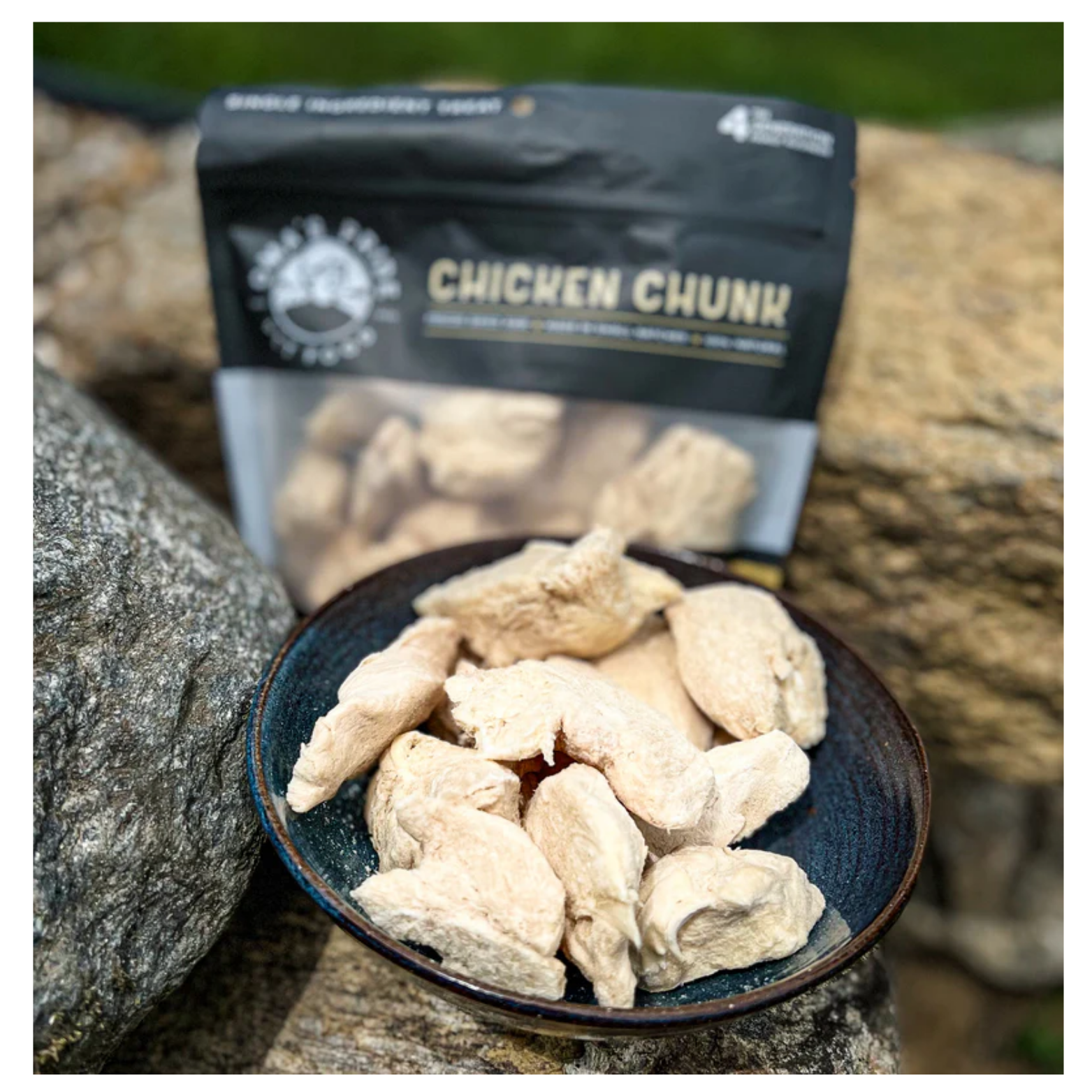 Freeze-dried chicken chunks in a ceramic bowl placed on rocks outdoors, with the Oma’s Pride Chicken Chunk treat bag visible behind the bowl.
