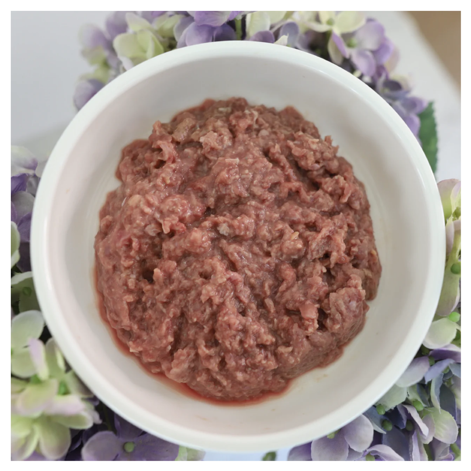A white bowl filled with raw ground poultry cat food sits centered, surrounded by decorative purple and white flowers in the background, emphasizing the texture of the food.