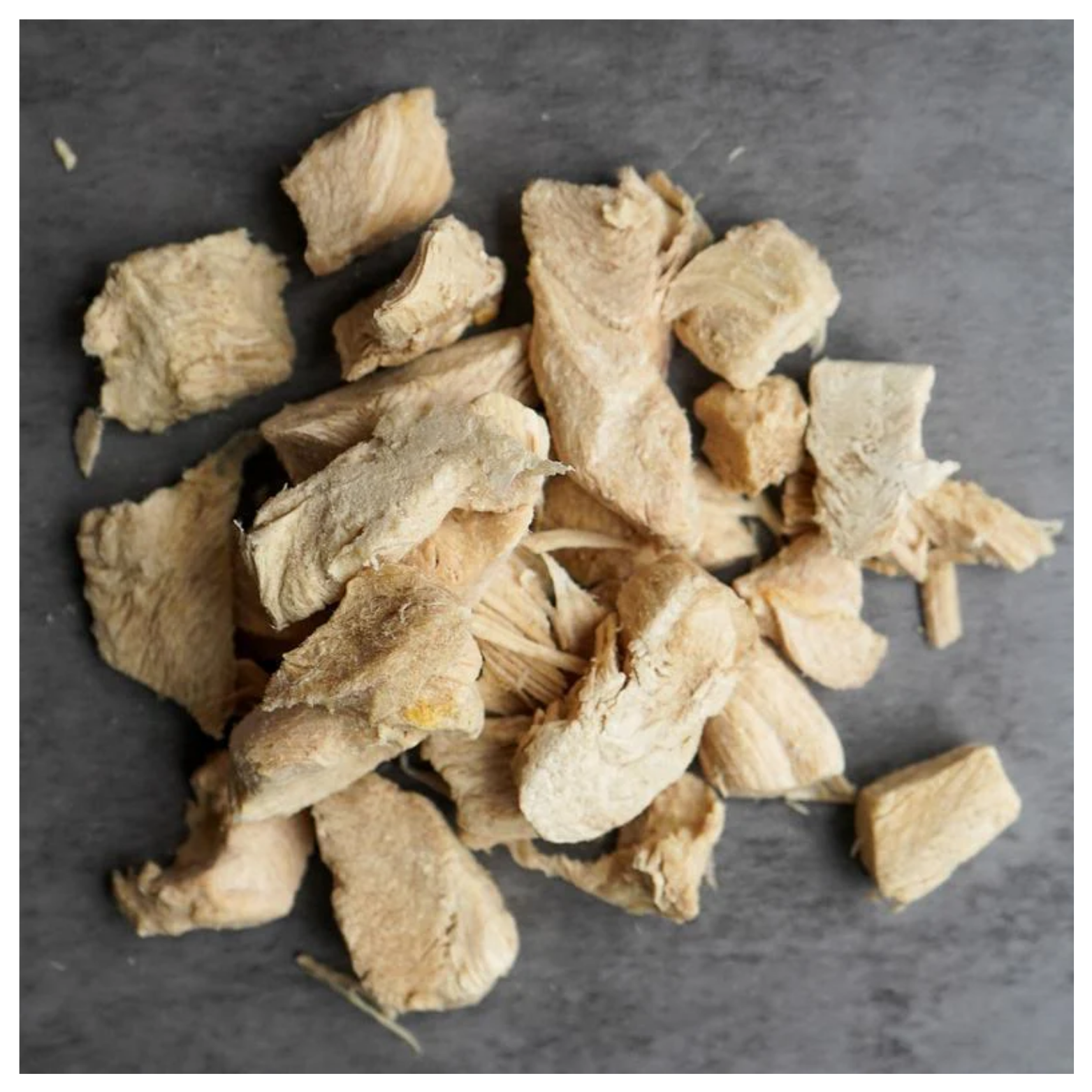 Close-up pile of irregular beige freeze-dried turkey breast chunks on a dark surface, showing fibrous texture and rough edges.