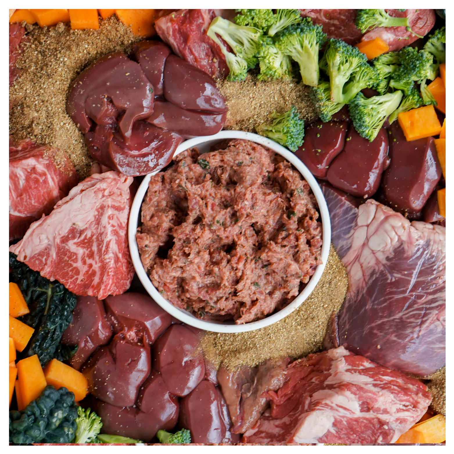 Bowl of ground raw beef dog food surrounded by raw beef cuts, organ meats, broccoli, kale, butternut squash, and a nutrient powder mixture arranged on a flat surface.