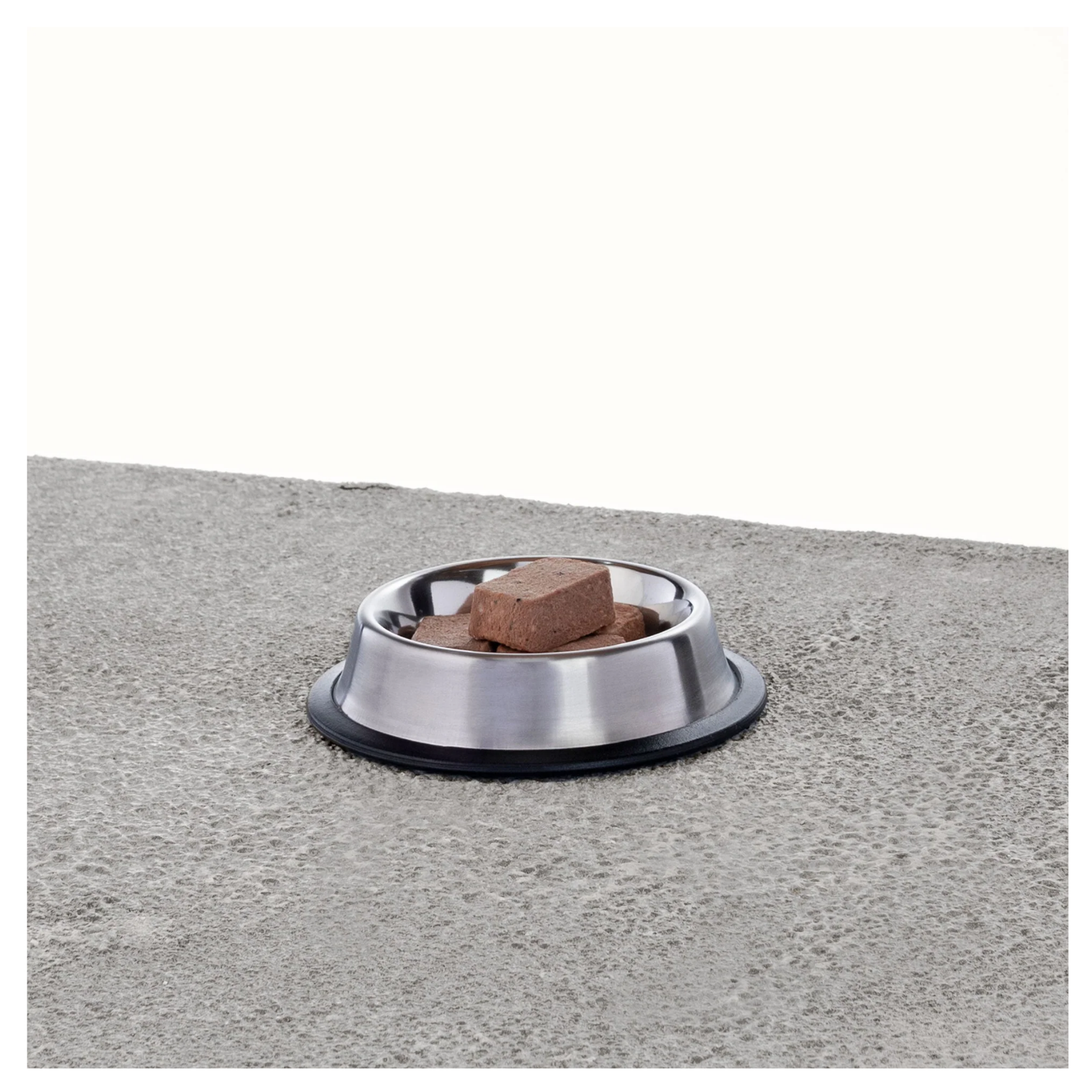 Stainless steel pet bowl containing several rectangular frozen raw nuggets placed on a textured gray surface with a white background.