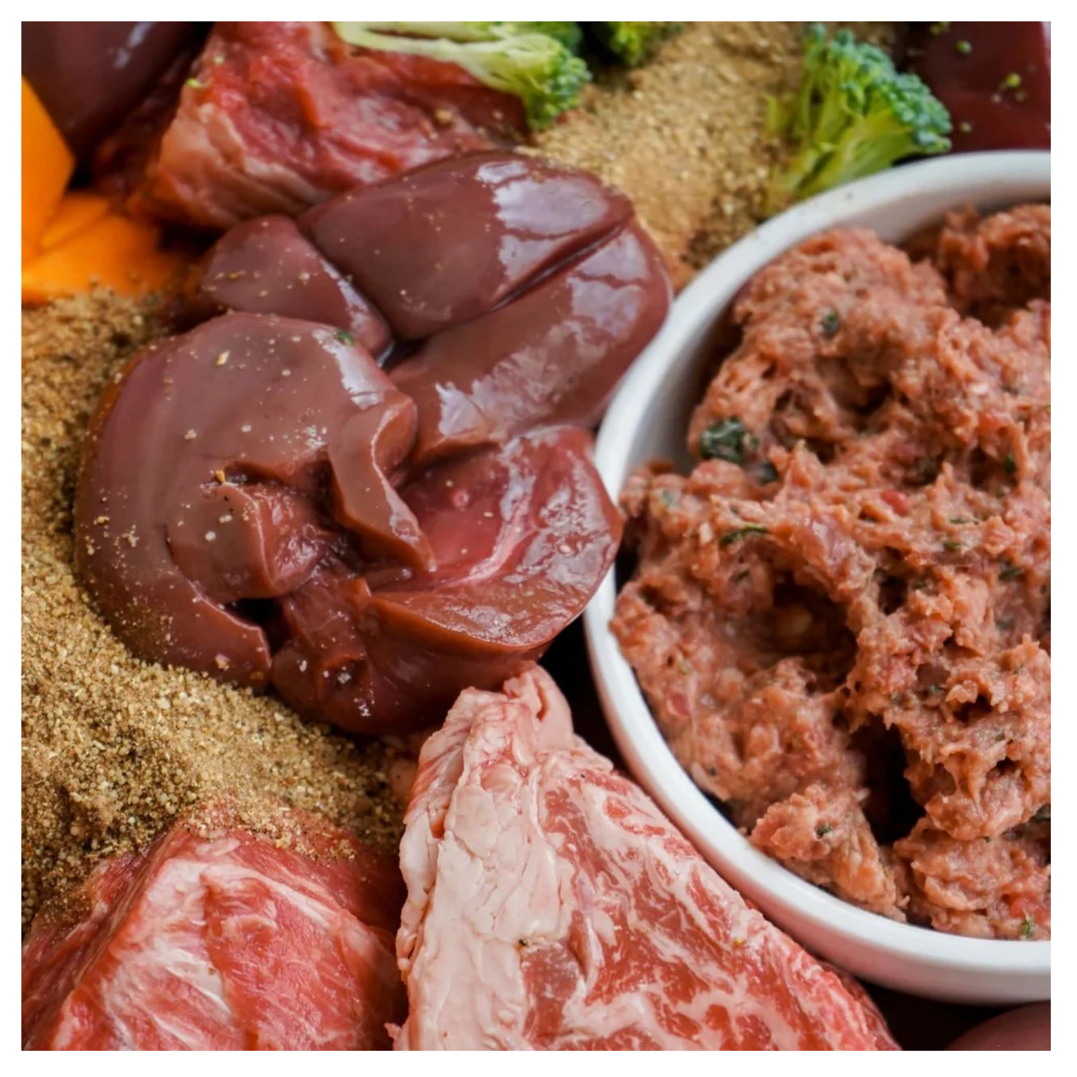 Close-up of raw beef ingredients including organ meats and nutrient powder next to a bowl of ground beef dog food.