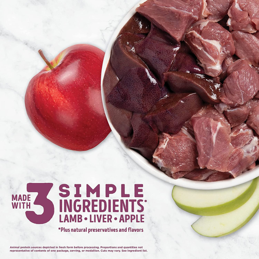 A top-down view showing the primary ingredients of the recipe. On the right, a white bowl is filled with raw chunks of red meat (lamb) and dark purple organ meat (liver). To the left, there is a whole shiny red apple and two green-skinned apple slices. Text in the corner reads: "MADE WITH 3 SIMPLE INGREDIENTS: LAMB • LIVER • APPLE."